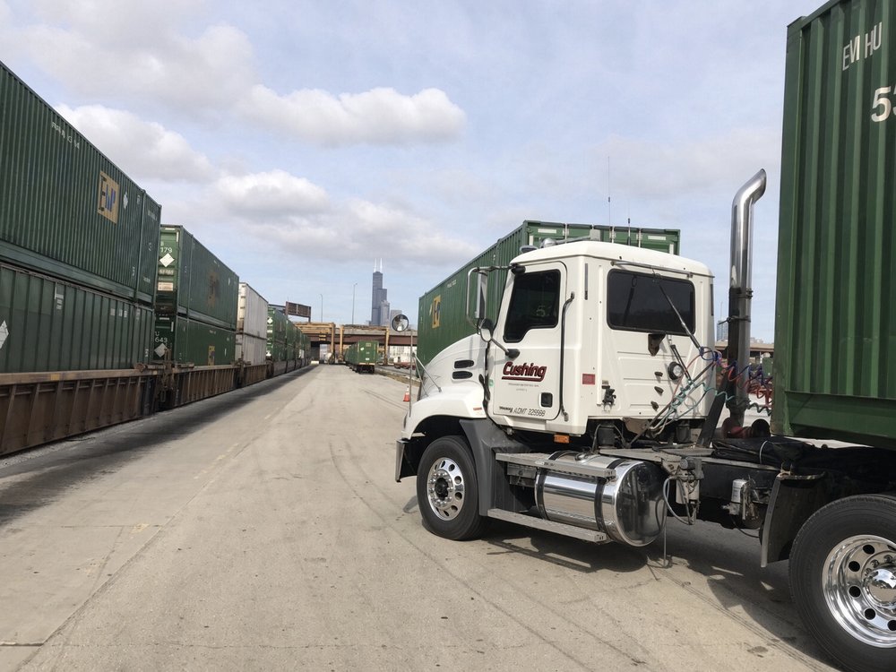 A white Cushing Mack day cab under a 53 foot container between two doublestack trains at UP Canal Street, Chicago, the Willis Tower on the horizon