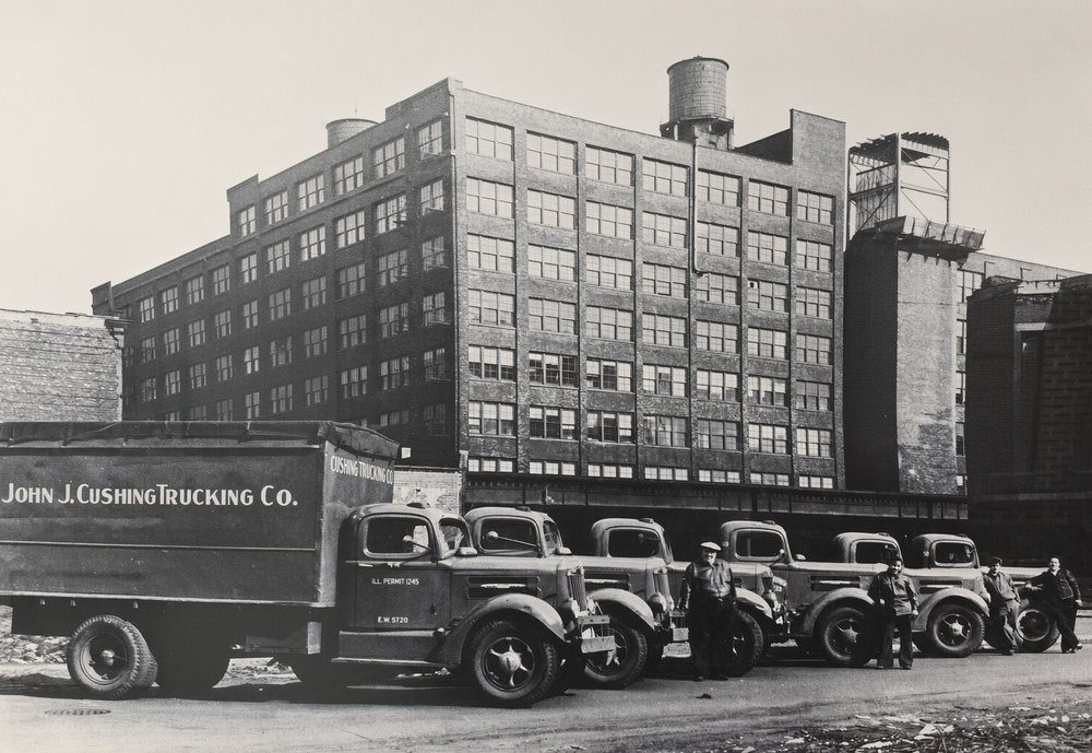A John J. Cushing Trucking Co. straight truck and a row of trucks with their drivers standing beside them, a tall brick warehouse behind