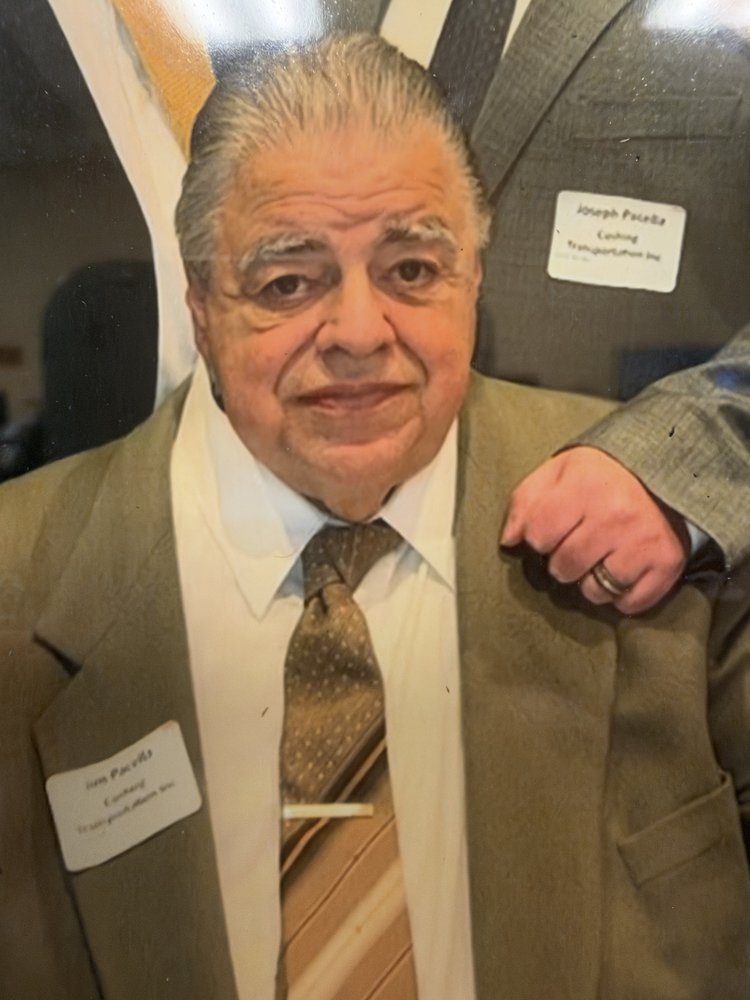 Jim Pacella in a tan suit, white shirt and patterned tie, a name badge on his lapel reading Jim Pacella, Cushing Transportation Inc; beside him a second badge reading Joseph Pacella, and a hand on his shoulder