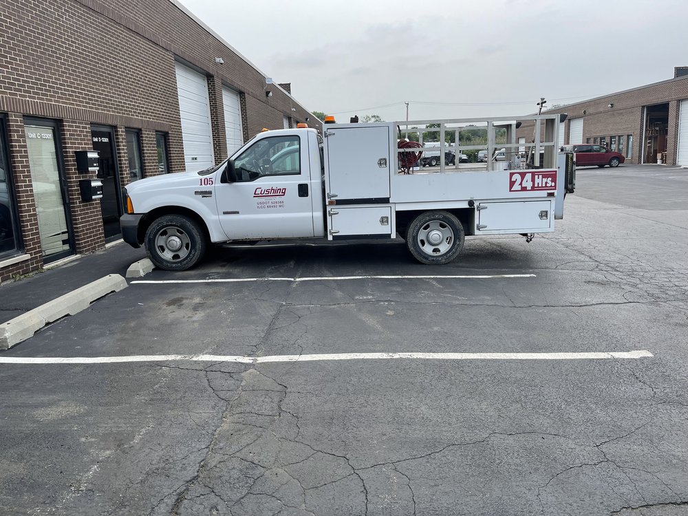 A white service truck lettered Cushing with a 24 Hrs sign on its body, parked at the brick office