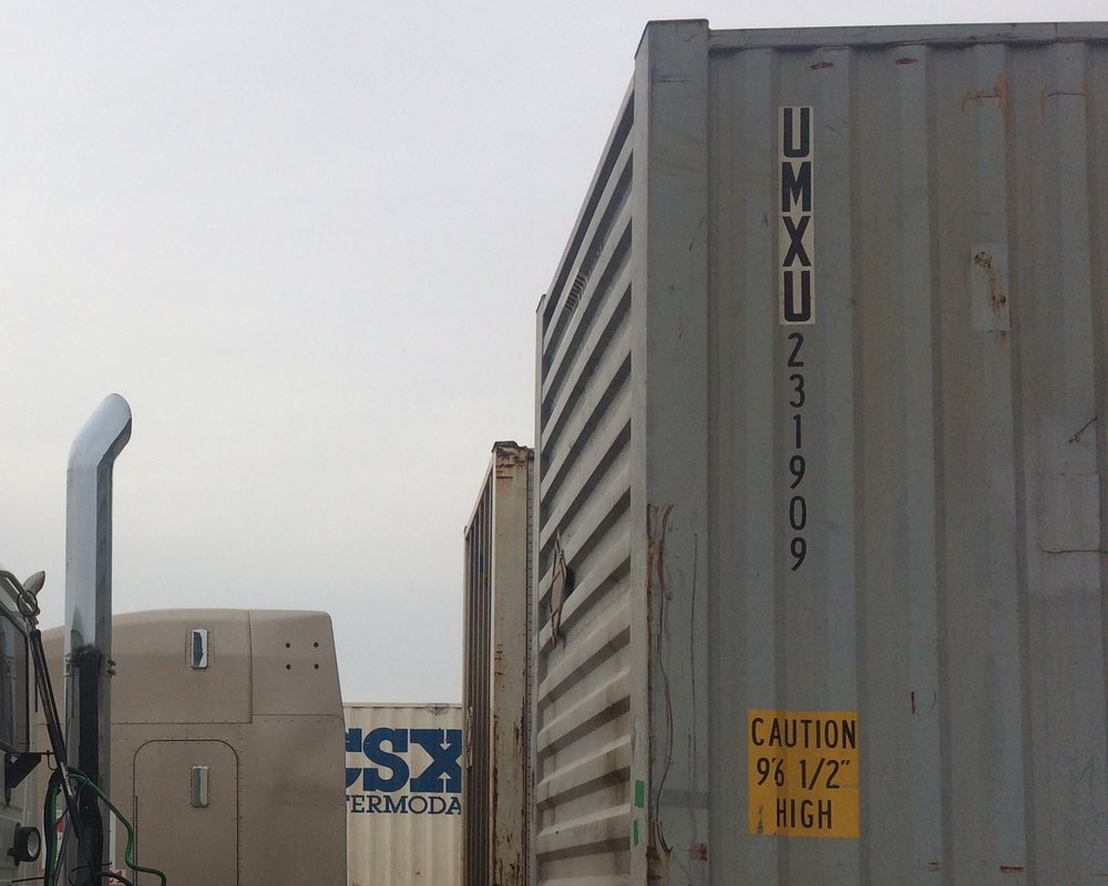 The corrugated grey side of a UMXU container on a Cushing chassis, its caution sign and lettering, a CSX Intermodal trailer and the back of a Cushing day cab behind, under an overcast sky