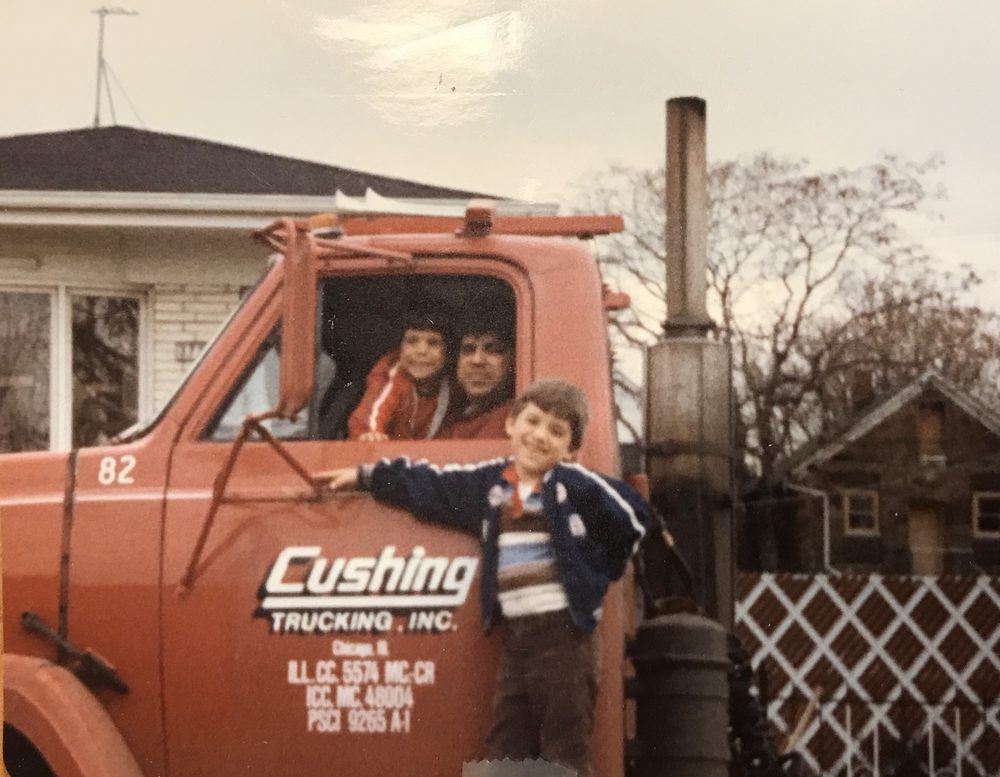 Jim Pacella at the window of red Cushing Trucking, Inc. unit 82 with a small boy on his lap, another boy on the step, a brick house and a lattice fence behind
